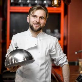 Chef cook portrait in uniform with steel cloche at the red restaurant kitchen interior. Image with small deph of field and blurred background